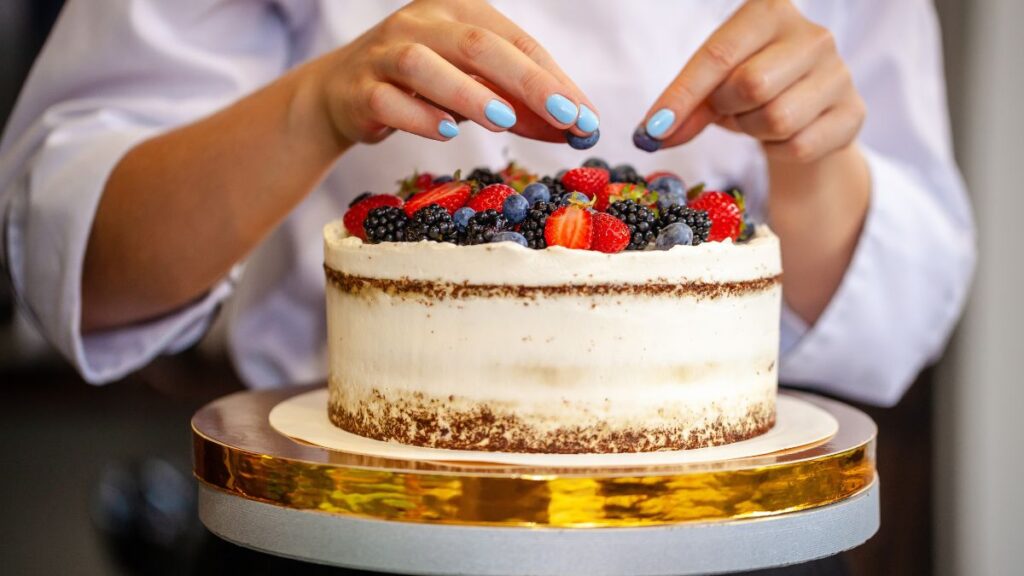 Hands decorating a frosted cake with fresh strawberries, blackberries, and blueberries on top.