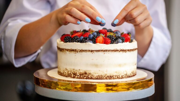 Hands decorating a frosted cake with fresh strawberries, blackberries, and blueberries on top.