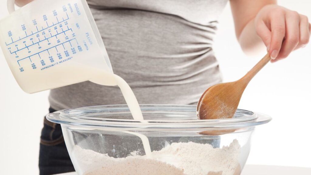 Pouring milk into a clear mixing bowl of dry cake mix with a wooden spoon.