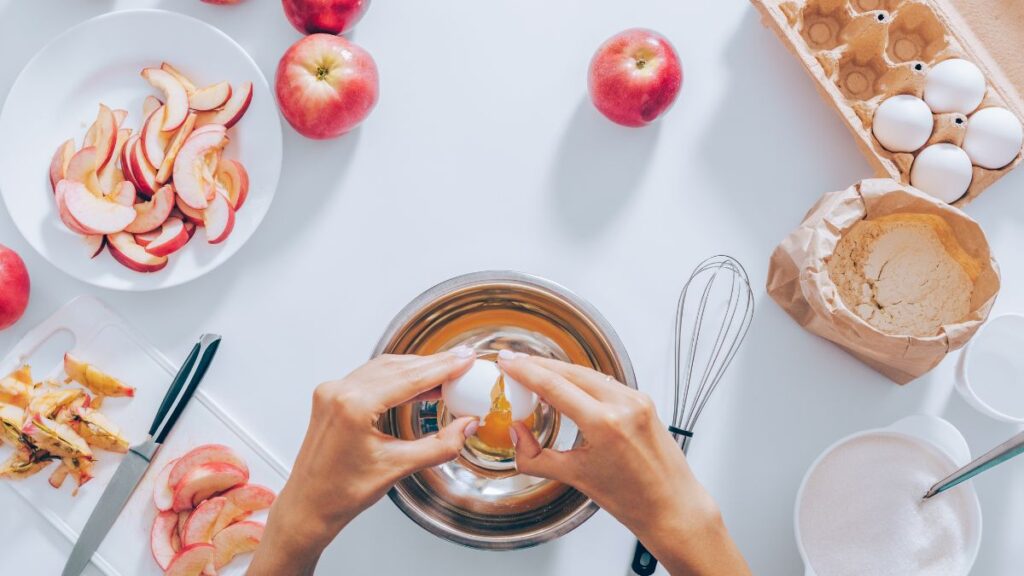 Cracking an egg into a mixing bowl surrounded by flour, sugar, and apples.