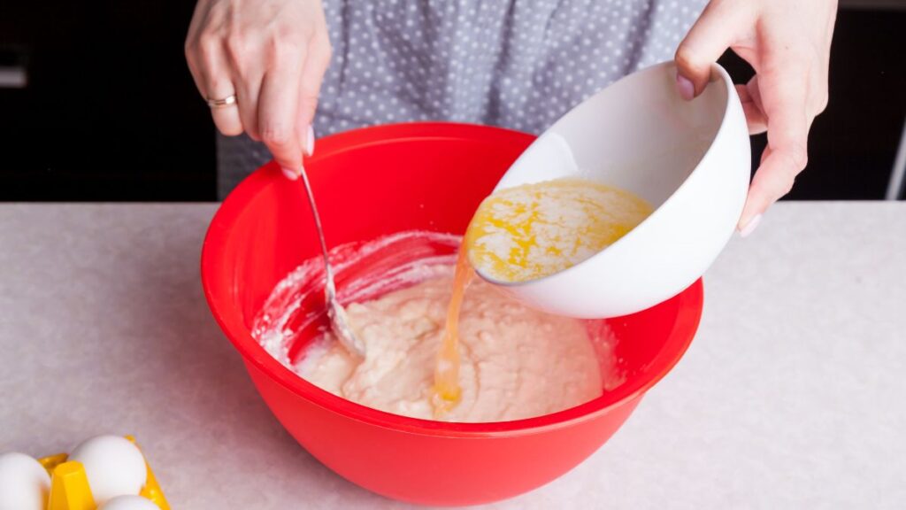 Adding melted butter into a red mixing bowl of boxed cake mix batter.