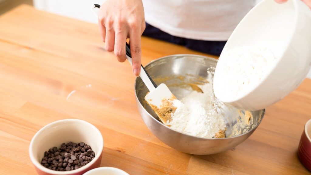 Mixing flour into cookie dough with a spatula next to a bowl of chocolate chips.