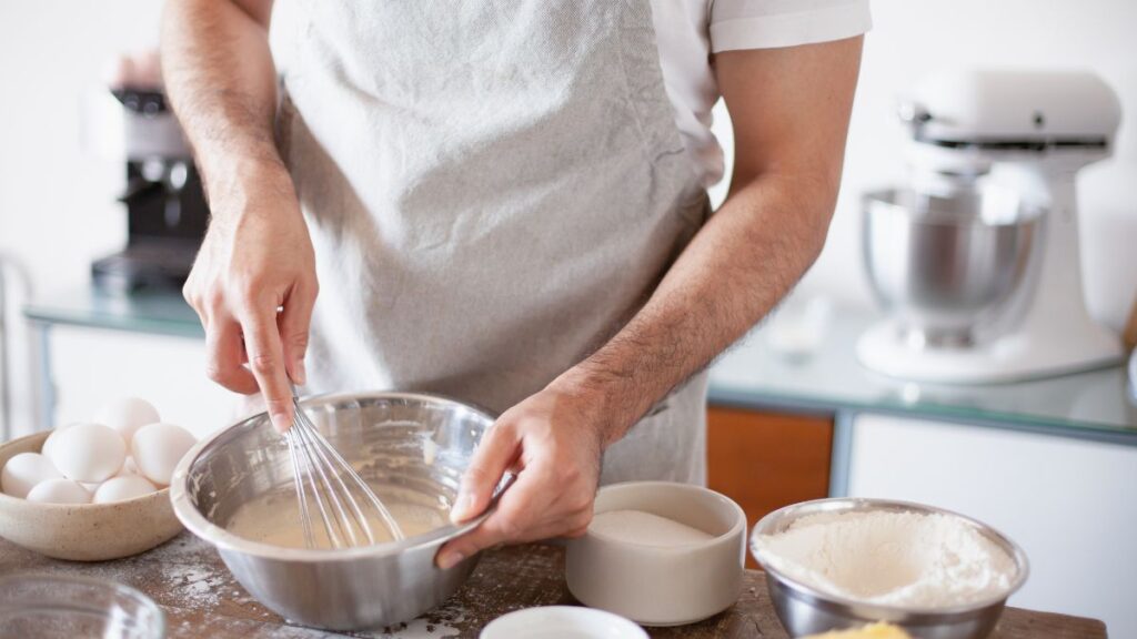 Whisking cake batter in a stainless steel bowl with eggs and flour nearby.