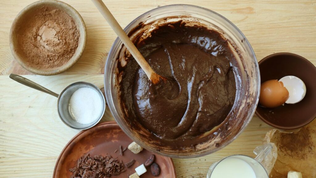 Thick chocolate cake batter in a bowl with cocoa powder, sugar, and eggshells on the side.