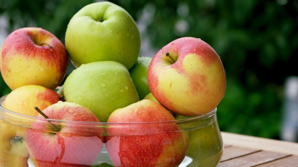 Glass bowl filled with fresh green and red apples outdoors.