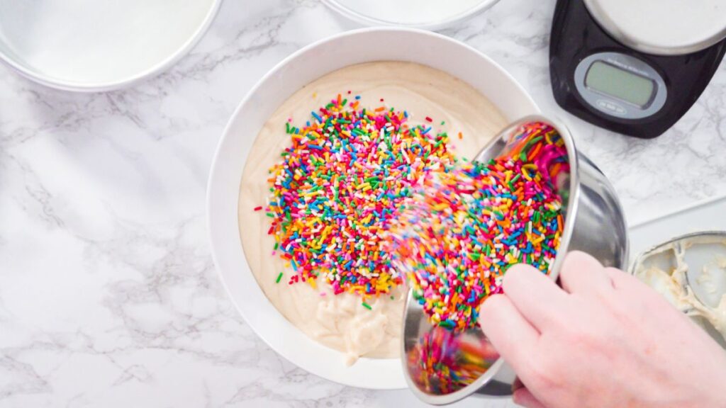 Pouring rainbow sprinkles into vanilla cake batter in a white bowl.