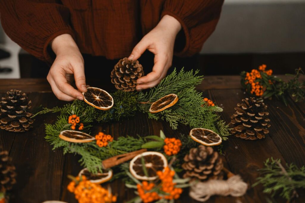 Hands creating a Christmas wreath with pinecones, dried oranges, and festive greenery.