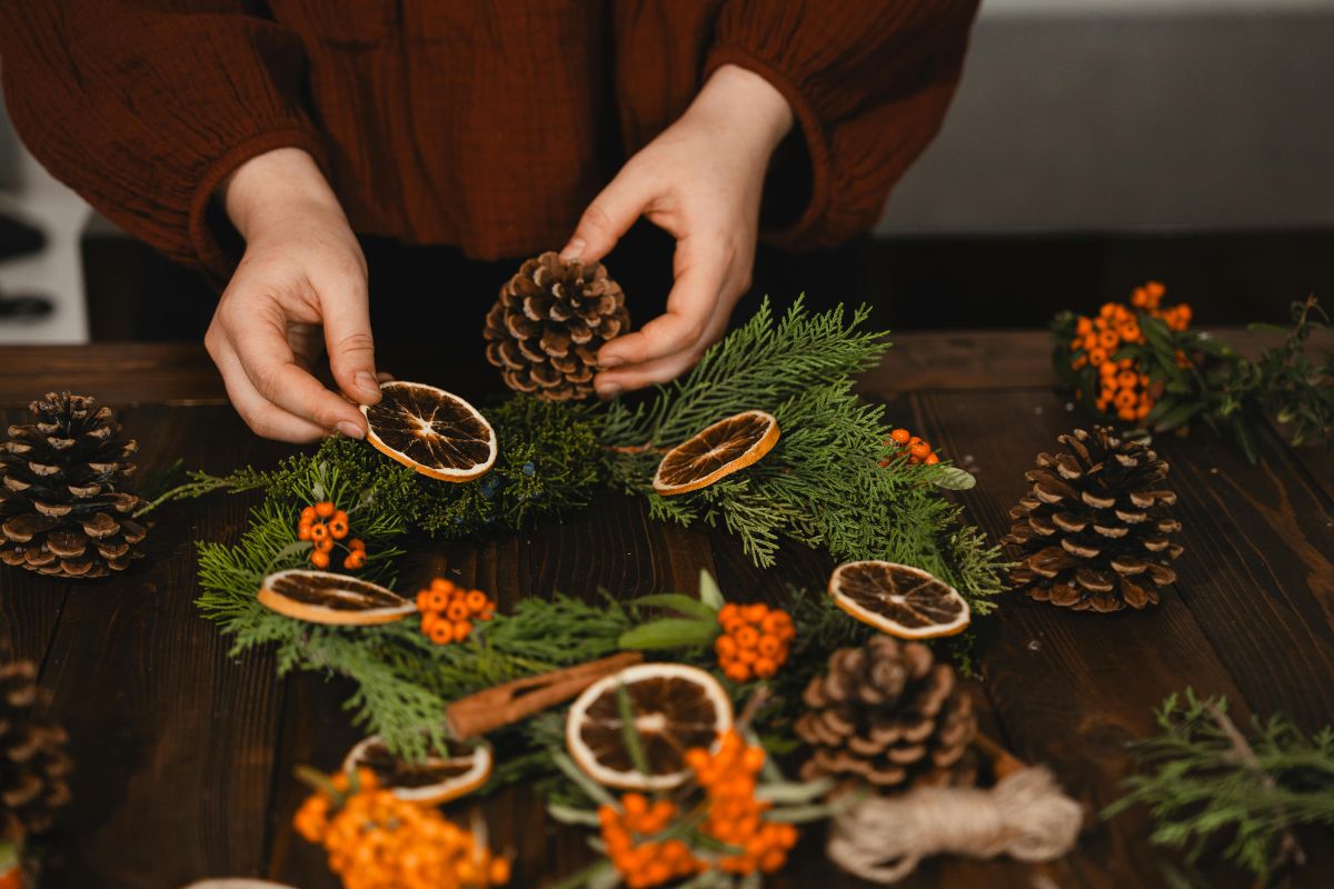 Hands creating a Christmas wreath with pinecones, dried oranges, and festive greenery.