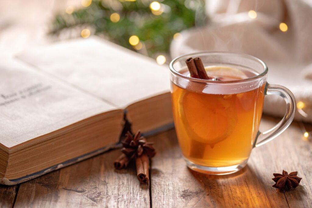 Glass mug of spiced tea with cinnamon and lemon beside an open book, a cozy Christmas self care moment.