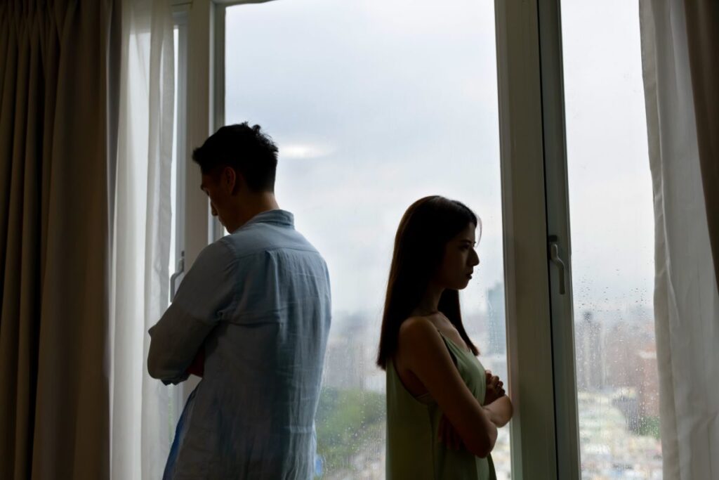 Couple standing apart in silence by a window, showing emotional distance.