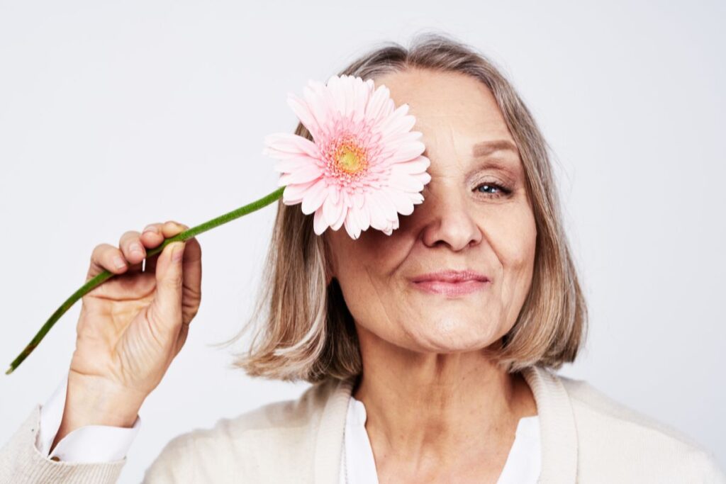 Confident older woman smiling with a pink flower covering one eye, showing boldness and empowerment.