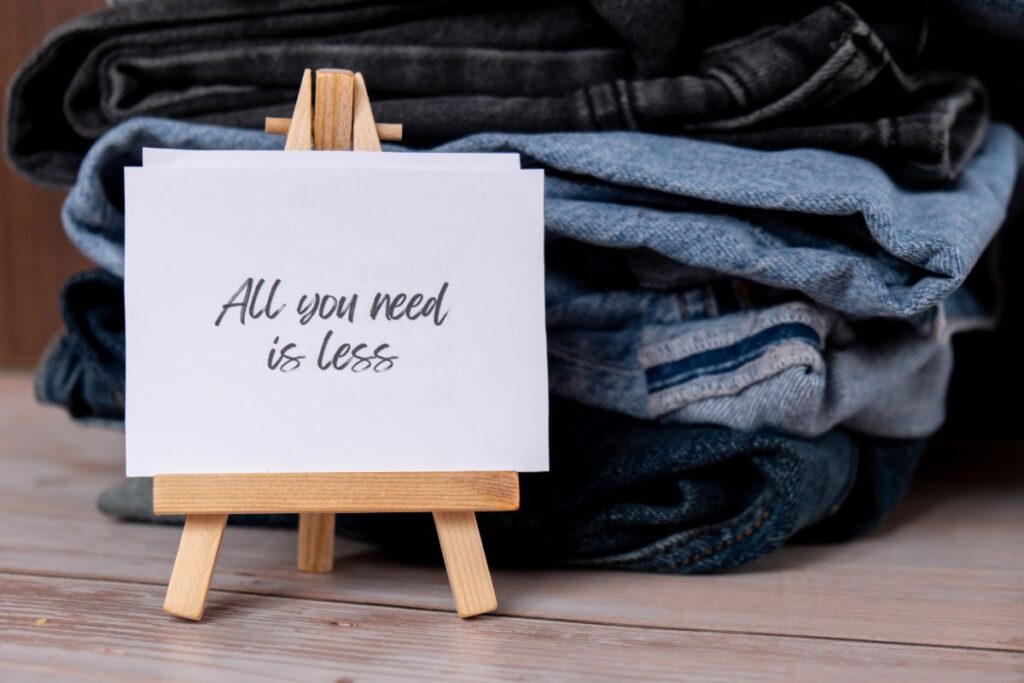 Sign saying all you need is less placed in front of folded jeans.