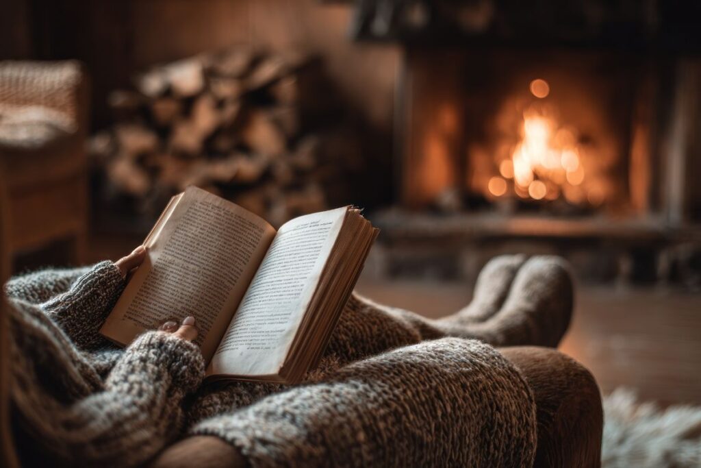 Person wrapped in blanket reading a book beside a glowing fireplace.