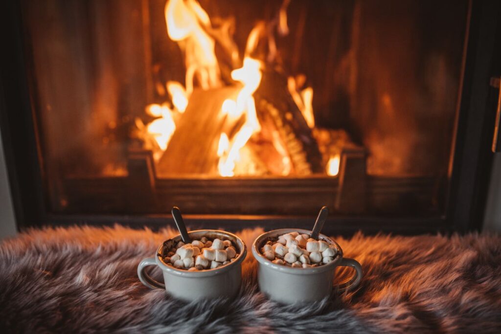 Two mugs of hot chocolate topped with marshmallows in front of a glowing fireplace.