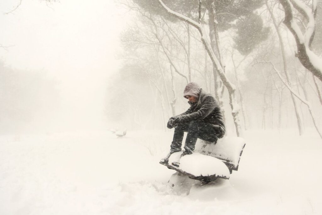 Person bundled in coat sitting on bench during heavy winter snowstorm.