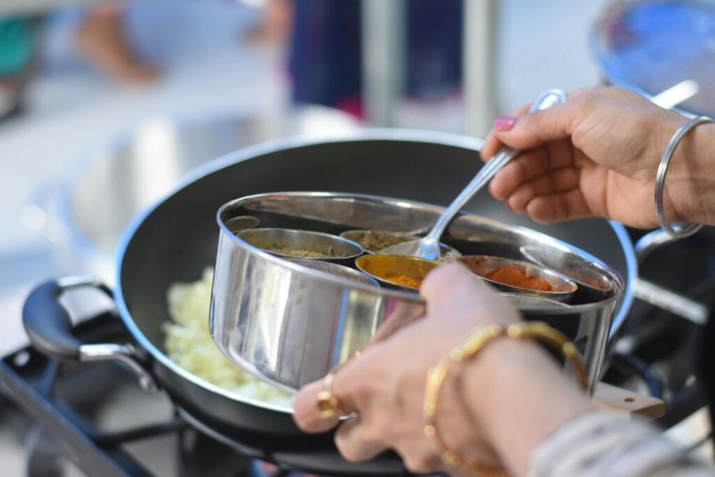 Hands holding a spice box over a stovetop while cooking in an Indian kitchen.
