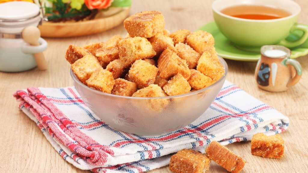 Bowl of golden brown homemade croutons on a checkered napkin beside a cup of tea.