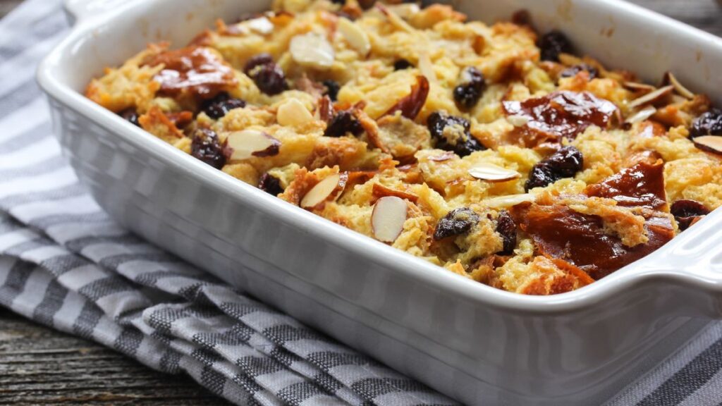 Baked bread pudding with raisins and almonds in a white ceramic dish on a striped towel.