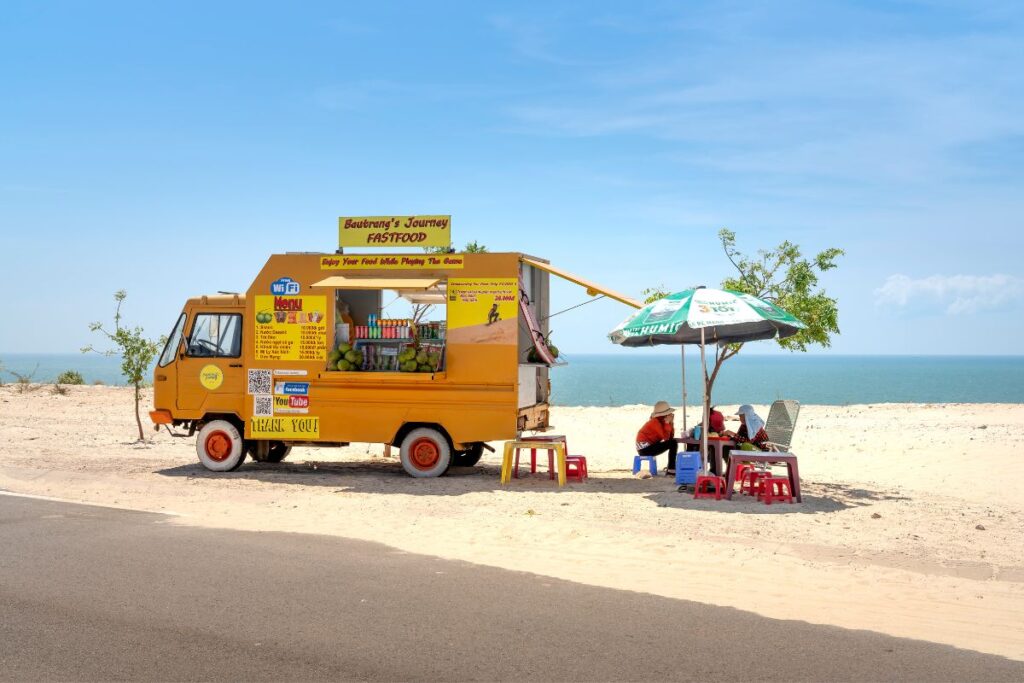 Bright yellow food truck parked near the beach serving snacks under an umbrella.