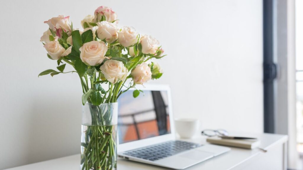 Soft pink roses in a vase placed near a laptop on a bright desk.