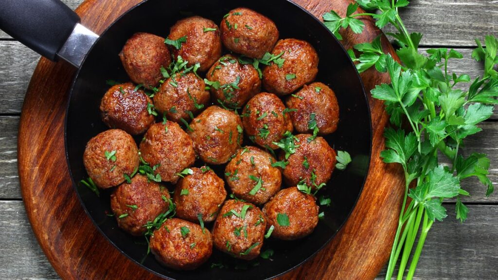 Skillet of browned meatballs garnished with chopped parsley on a wooden board.