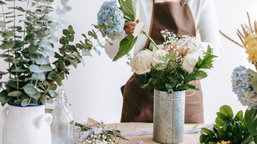 Florist arranging white and blue hydrangeas in a metal vase surrounded by greenery.