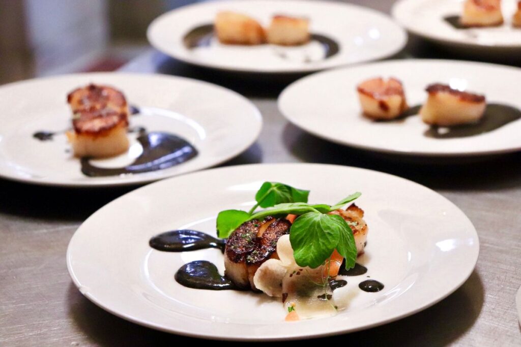 Gourmet scallops plated on white dishes with dark sauce and fresh greens.