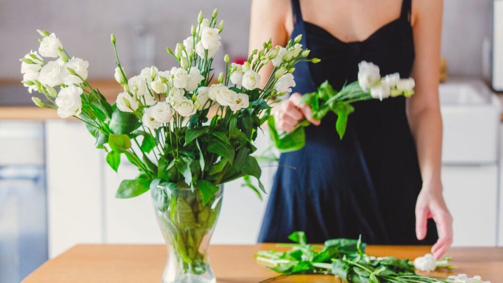 Woman removing wilted stems from a vase of white lisianthus flowers on a kitchen counter.