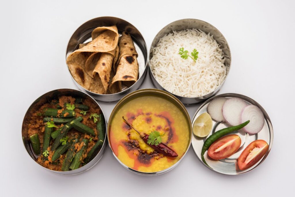 Traditional Indian meal in lunch box with rice, dal, chapati, okra curry, and fresh sides.