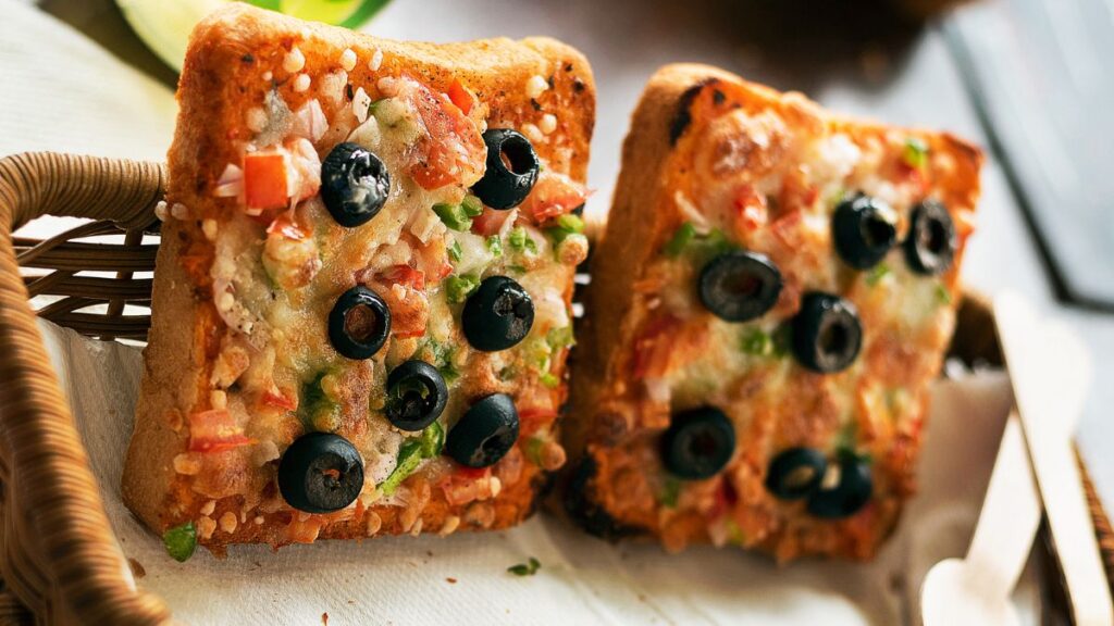 Toasted bread pizzas topped with cheese, black olives, and diced vegetables in a basket.