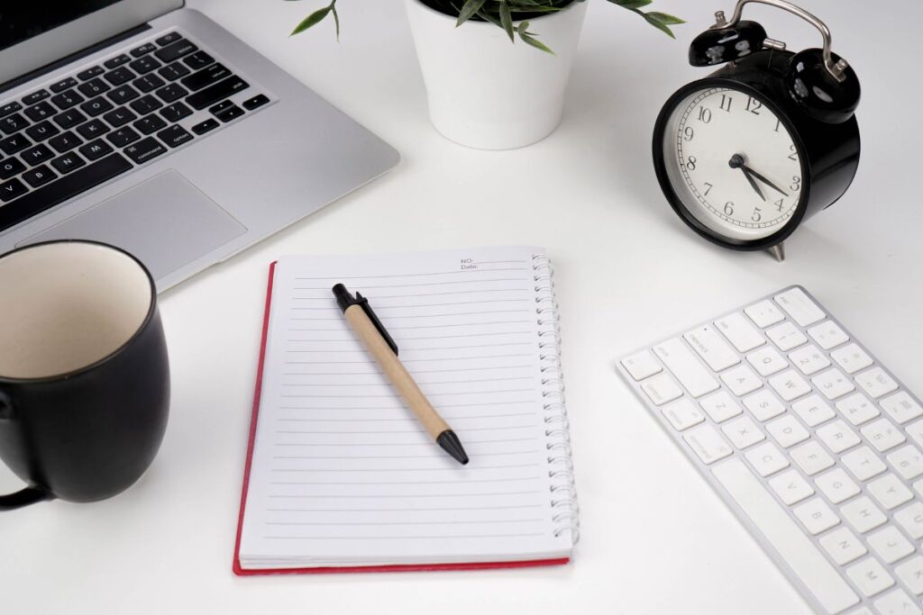 Notebook with pen, coffee, clock, and laptop for work planning.