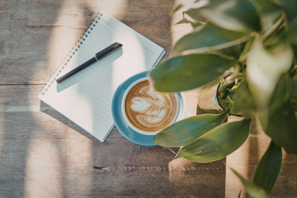 Notebook and pen beside a latte on a wooden table with sunlight and plants.
