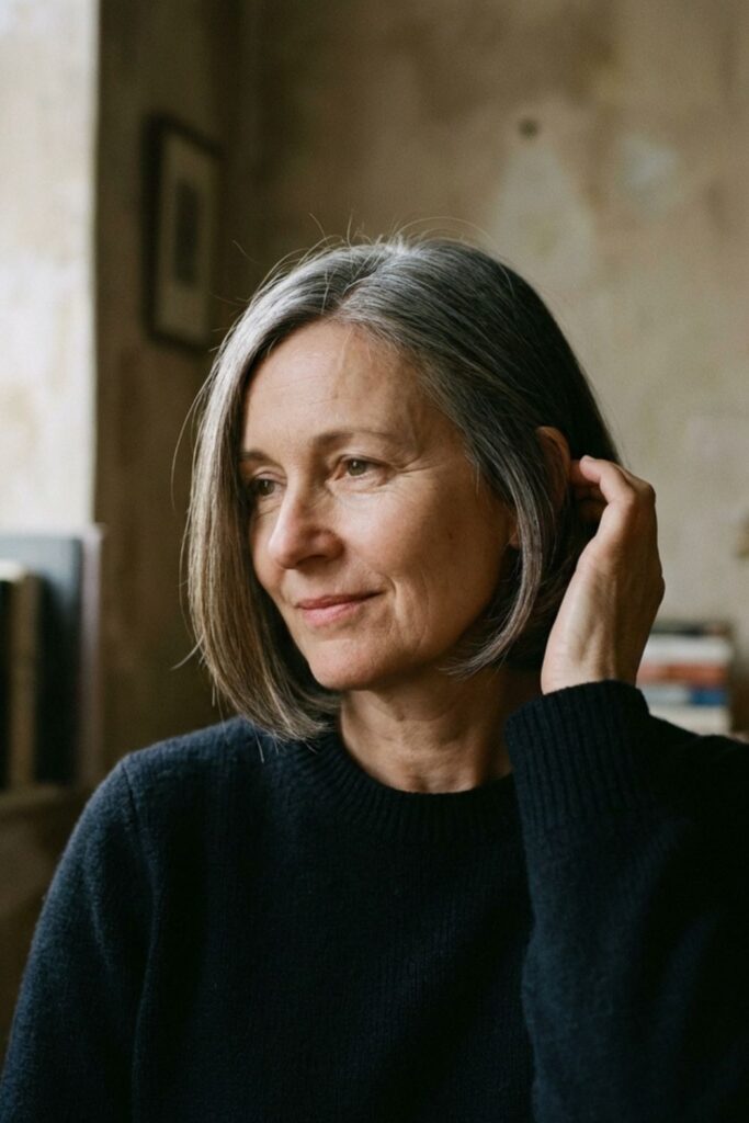 Woman with a sleek gray bob sitting indoors near bookshelves, gently touching her hair and looking to the side.