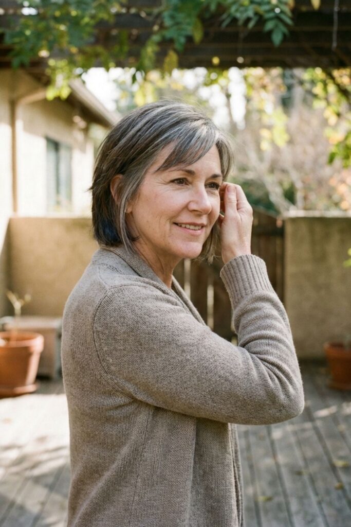 Woman in a knit sweater standing on a wooden patio surrounded by greenery, touching her hair.