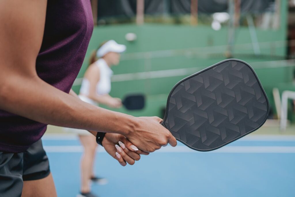 Close-up of a woman holding a black pickleball paddle, dressed in sporty chic activewear on an outdoor court.