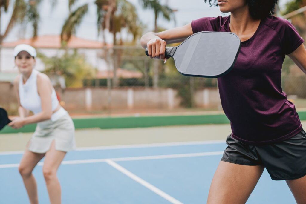 Two women playing pickleball in stylish court outfits, wearing sporty tops and skorts on a sunny outdoor court.