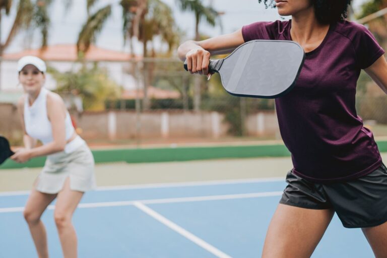 Two women playing pickleball in stylish court outfits, wearing sporty tops and skorts on a sunny outdoor court.