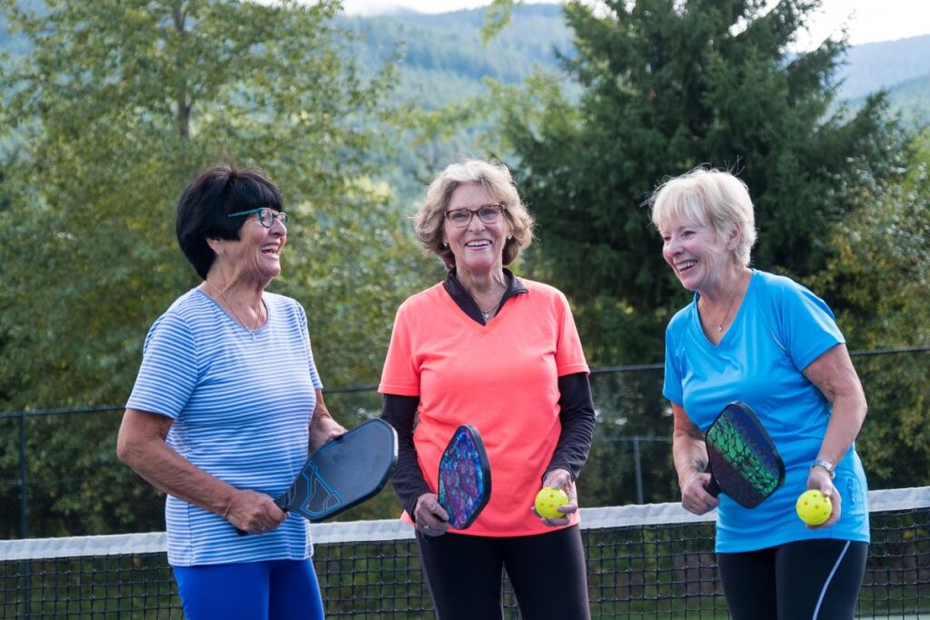 Three women over 50 laughing together on a pickleball court in colorful activewear holding paddles and balls.