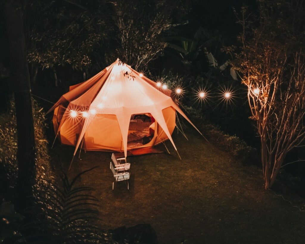 A circular, canvas bell tent glowing with warm orange light from inside and decorated with string lights draped over the entrance, set up at night on a dark grassy lawn surrounded by trees and foliage. A small white outdoor chair sits in front of the tent entrance.