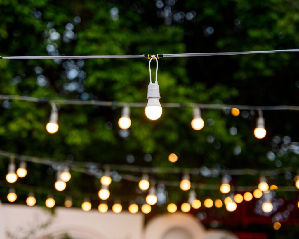A close-up of a string of warm white LED patio lights hanging outdoors against a dark green, out-of-focus background of trees. One light bulb is in sharp focus, while the rest create a soft bokeh effect.