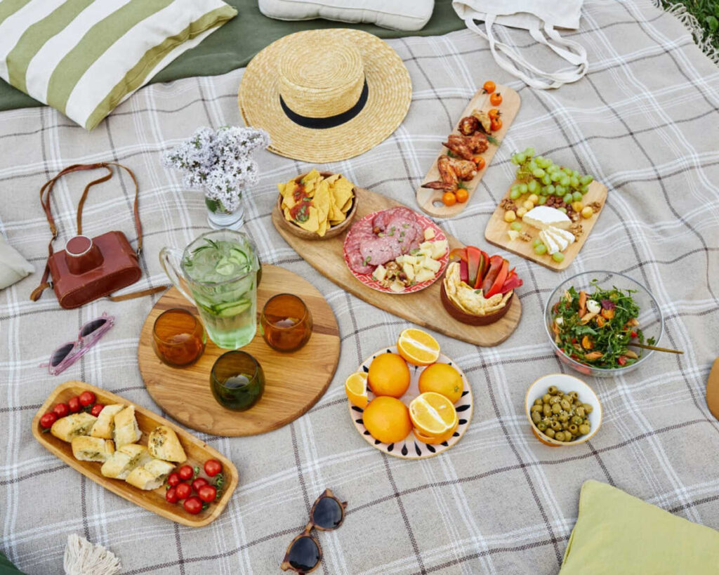An overhead view of a styled picnic blanket spread with an assortment of food and accessories. The food includes charcuterie boards with cured meat, cheese, and grapes, a bowl of olives, a side salad, and fresh oranges, along with drinks garnished with cucumber. Accessories include a straw hat, sunglasses, a leather camera bag, and striped pillows.