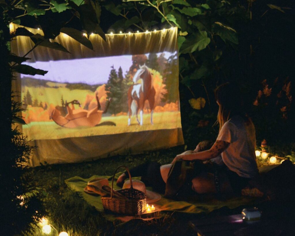 A cozy outdoor movie night in a backyard at dark, featuring a projected animated film of horses on a screen hung from trees. A person sits on a blanket in the foreground with a woven picnic basket, surrounded by warm string lights and lit candles on the ground.