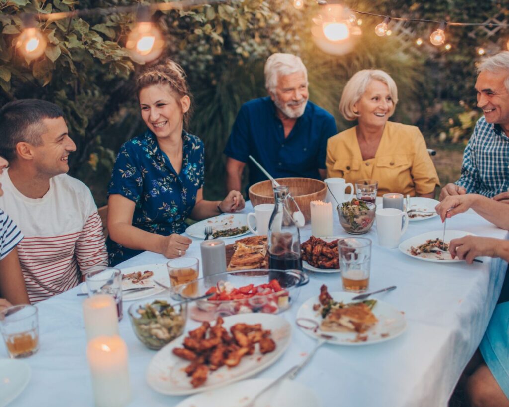A diverse group of family members or friends, including younger and older adults, smiling and conversing while gathered around a white-linen covered table for an outdoor dinner party at dusk. The table is filled with plates of food, wine, and lit candles, with warm string lights hanging overhead from the surrounding trees.