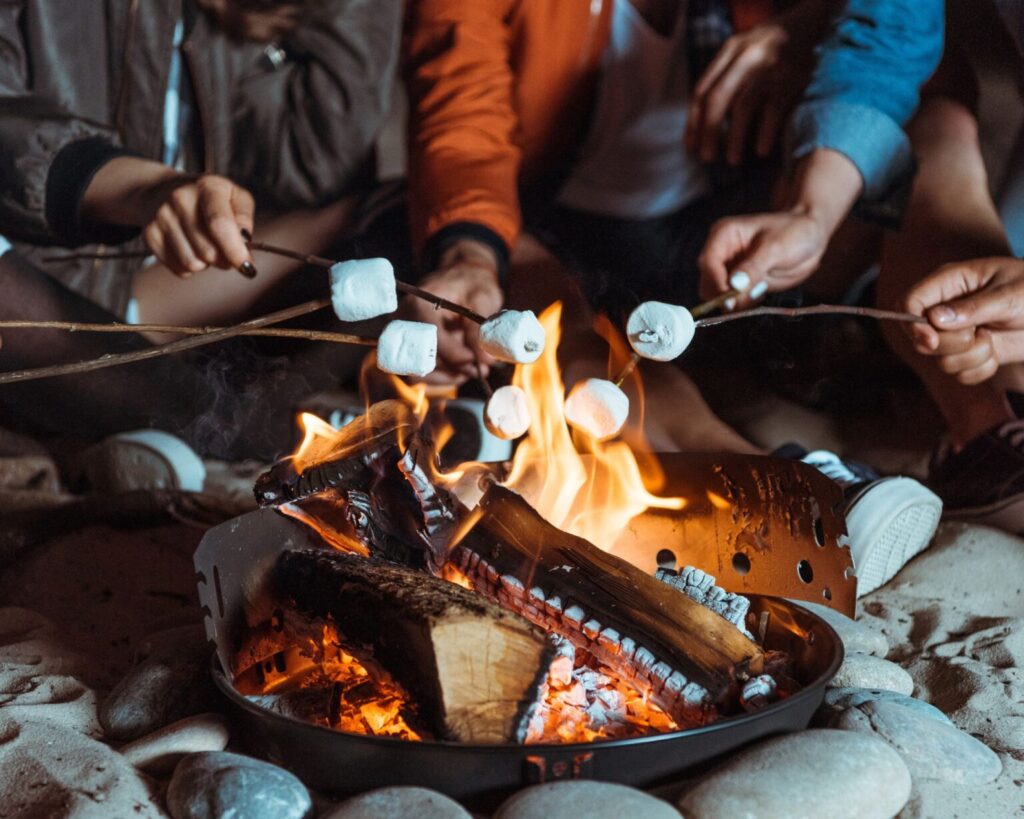 A close-up of a group of people sitting around a metal fire pit on the sand or ground at night, holding wooden sticks with marshmallows over the flames for roasting. Only their hands, shoes, and clothing are visible around the warm, glowing fire.