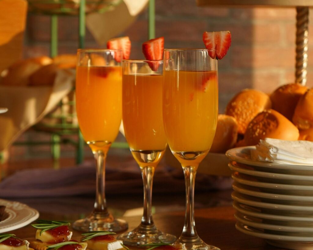 Three champagne flutes filled with a bright orange cocktail, such as a mimosa, each garnished with a sliced fresh strawberry on the rim. The drinks are set on a table with a stack of plates and a basket of bread rolls or pastries in the warm sunlight.