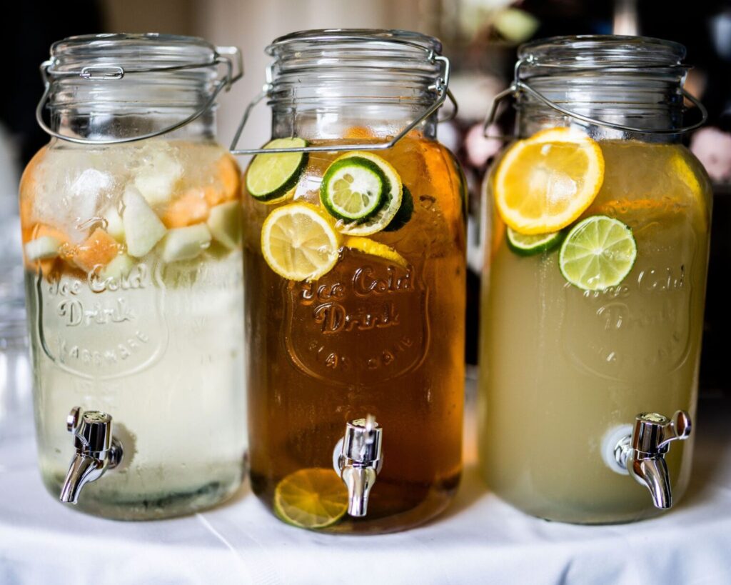 Three large glass mason jar beverage dispensers lined up on a table, each filled with a different infused drink for a party. The drinks include one with light-colored fruit (like apples or pears), one with iced tea and lime/lemon slices, and one with lemonade garnished with orange and lime slices.
