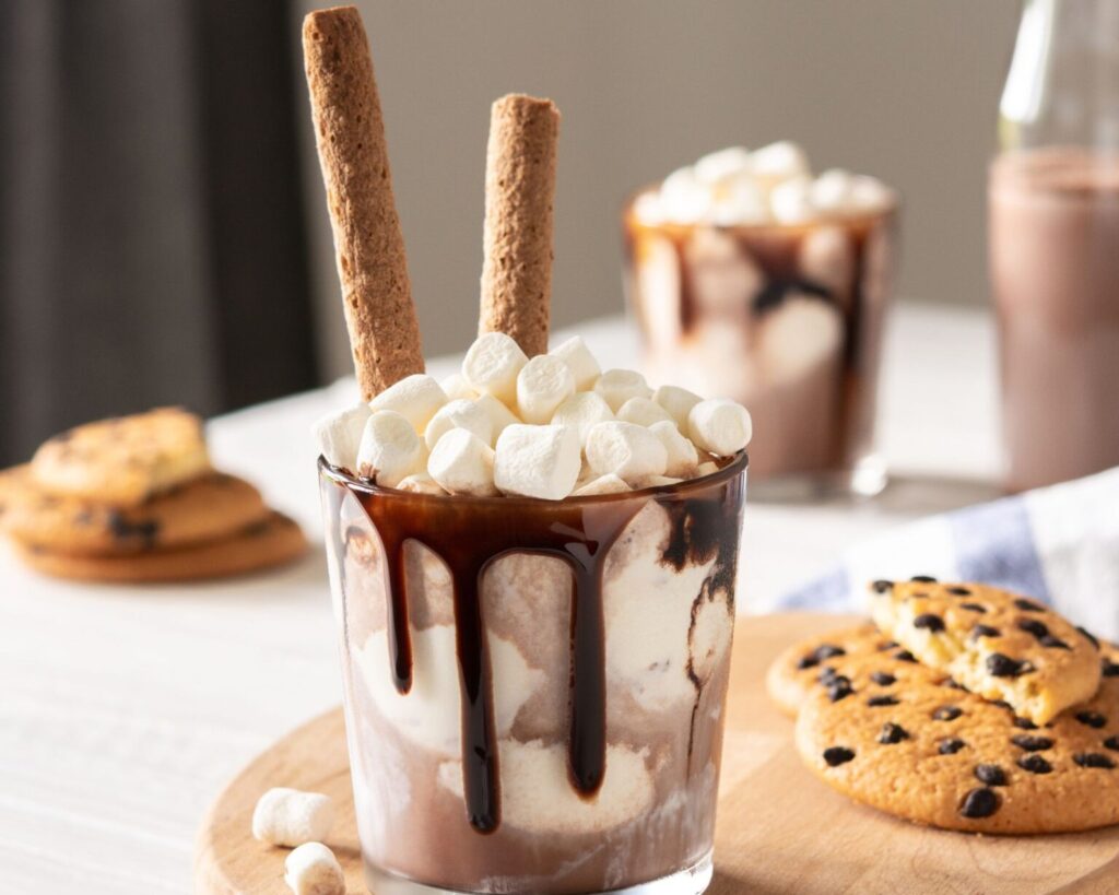 A glass mug filled with a chocolate milkshake or cold hot chocolate, drizzled heavily with chocolate syrup and piled high with mini marshmallows and two wafer sticks. The glass is set on a wooden cutting board with chocolate chip cookies nearby.