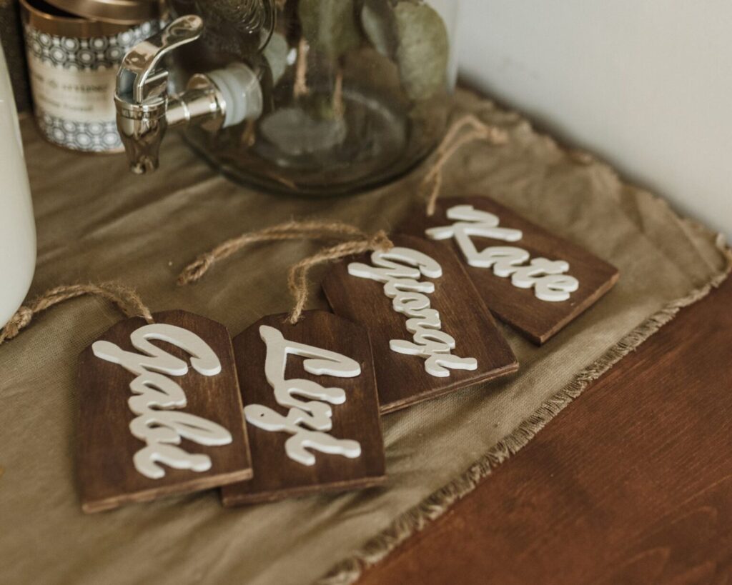 Four dark wooden, tag-shaped drink markers or place cards, each displaying a name (Gabi, Liz, George, Kate) in raised white script lettering. The tags are tied with twine and are resting on a sage green linen runner on a wooden surface, next to a glass beverage dispenser spigot.