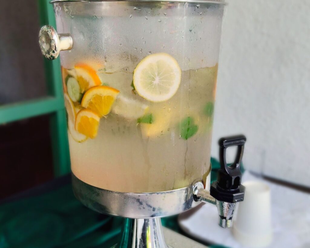 A clear, condensation-covered glass beverage dispenser filled with light-colored infused water or lemonade, containing slices of lemon, orange, and fresh mint leaves, set on a dark green tablecloth with a black spigot.