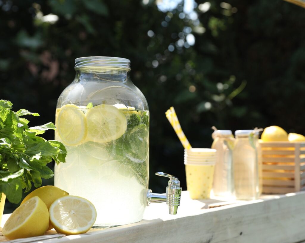 A large clear glass beverage dispenser filled with lemonade or infused water, containing lemon slices and mint leaves. It is set on a light wooden surface outdoors next to fresh lemons, a bunch of mint, yellow paper cups, glass bottles, and a wooden crate of more lemons, all against a background of dark green foliage.